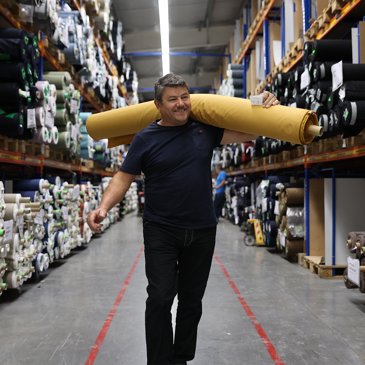 Man carrying a large yellow roll of fabric in a warehouse.